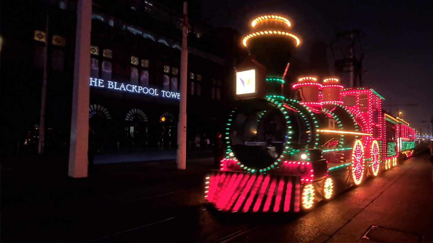 An illuminated steam train passes by the entrance to The Blackpool Tower